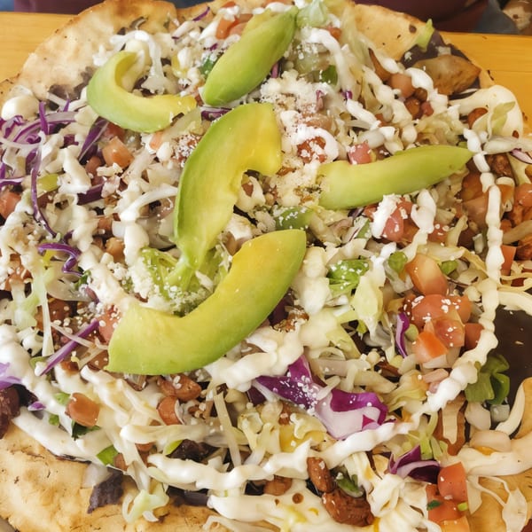 Close-up of a large El Azteca Mexican tostada topped with avocado, lettuce, cheese, and crema.